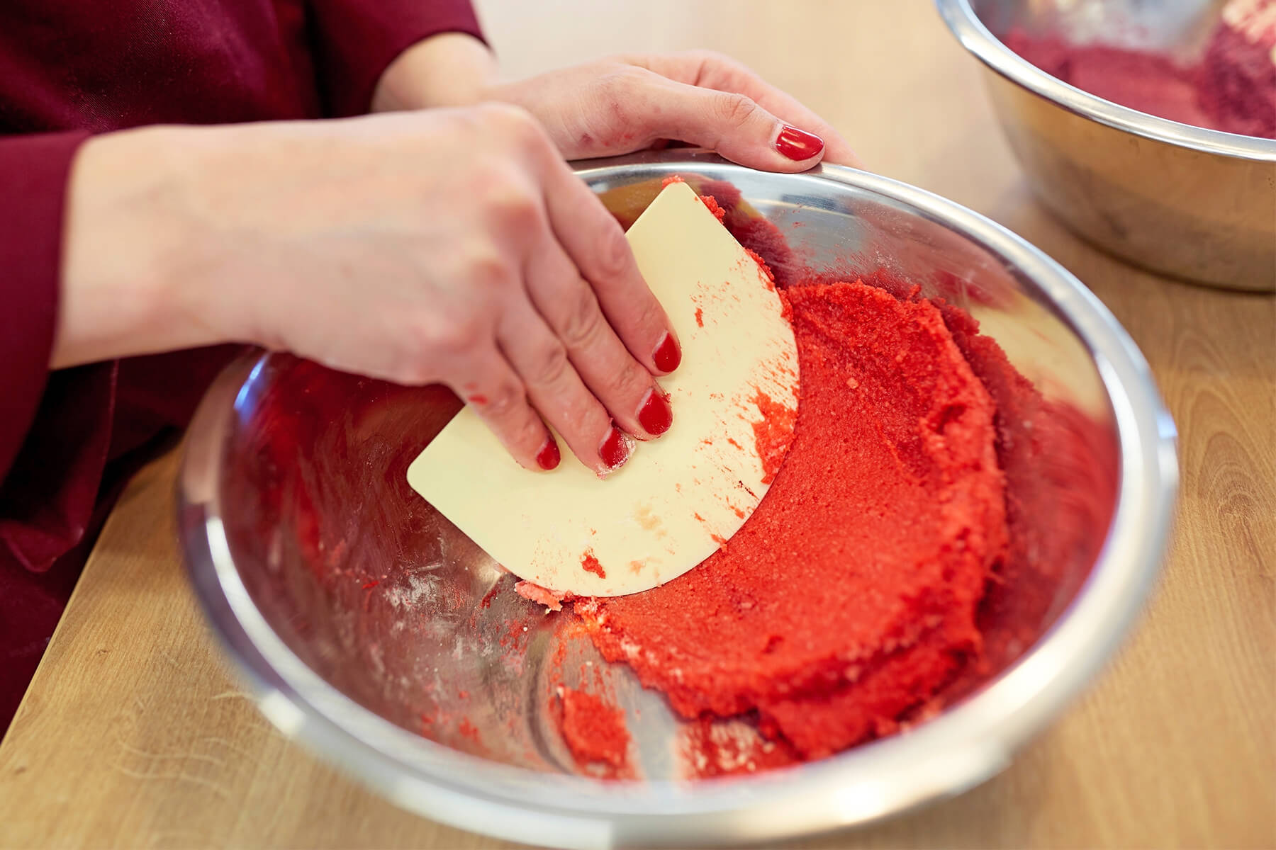 Eine kompakte rote Mandelmasse liegt als schwerer Klumpen in einer Edelstahlschüssel, eine Hand hält eine helle Teigkarte darüber