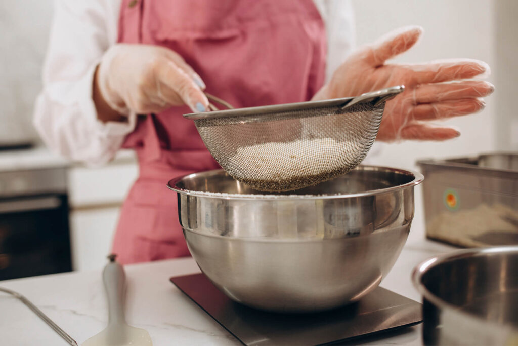 Eine Hand streicht eine helle Mischung aus gemahlenen Mandeln und Puderzucker durch ein rundes Sieb in eine Edelstahlschüssel, die auf einer Küchenwaage steht