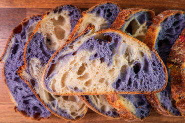 Scheiben eines Sauerteigbrots mit goldbrauner Kruste, in denen sich helle und violette Krume in breiten Bändern ineinanderwinden, auf einem Holzbrett