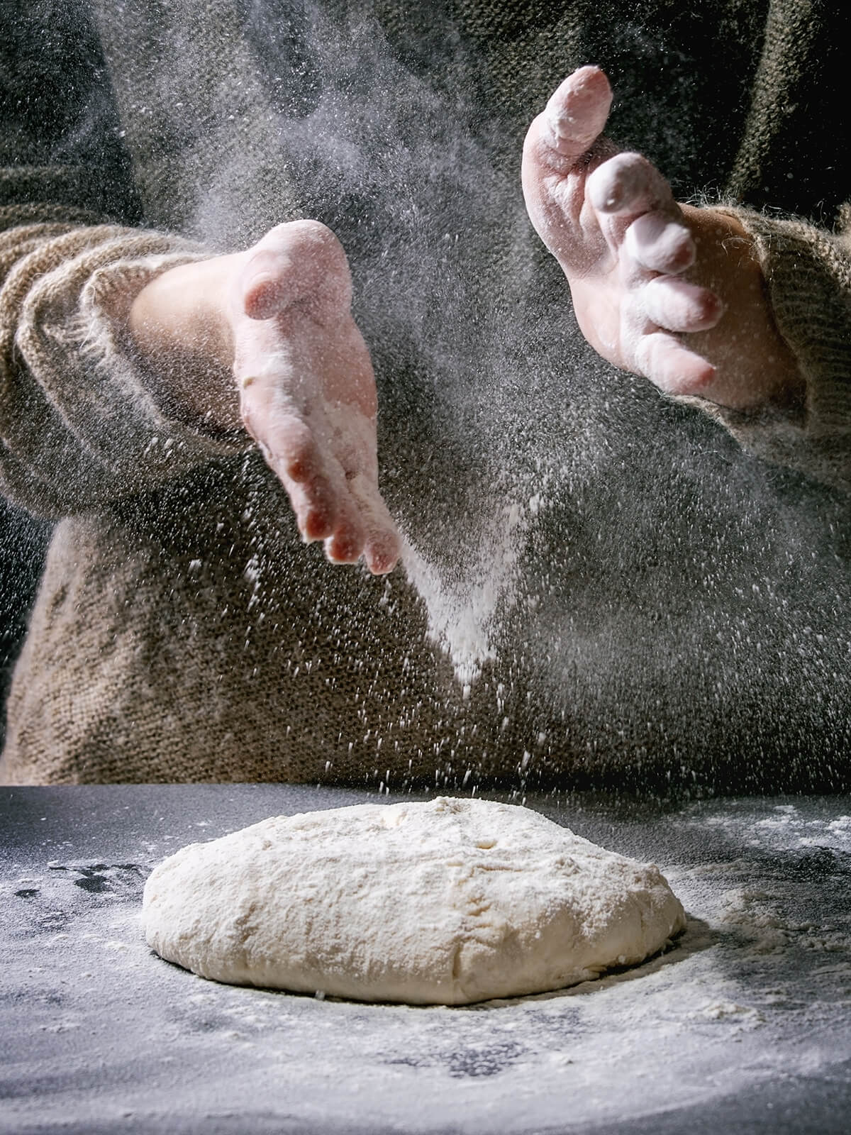 Mehl rieselt aus zwei bemehlten Händen auf einen flachen, weichen Brotteig auf einer dunklen Arbeitsfläche