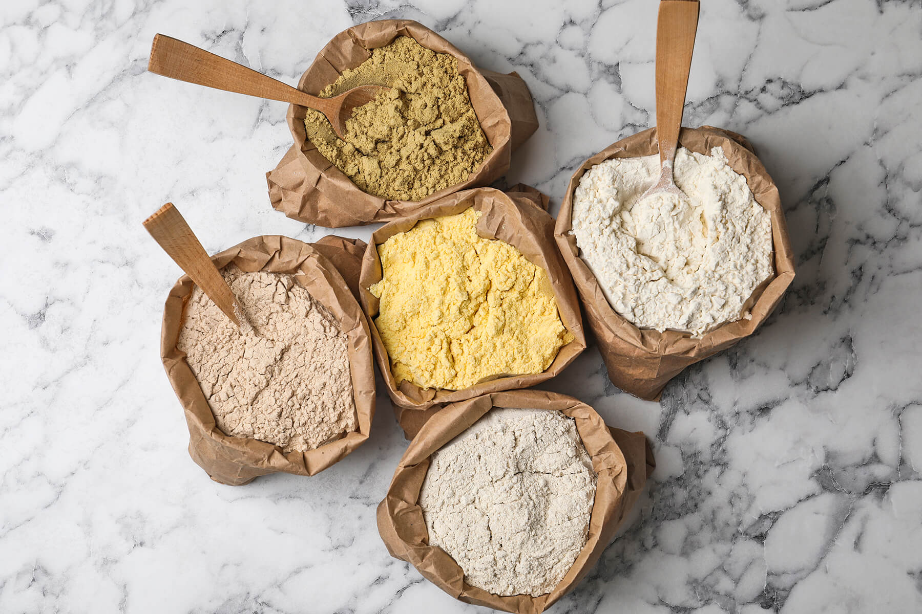 Fünf offene Papiersäcke mit grünlichem, gelbem, weißem, rosabeigem und graubeigem Mehl auf weißem Marmor, dazu ein Holzlöffel und zwei flache Holzschaufeln