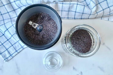 Geöffnete schwarze elektrische Kaffeemühle mit gemahlenem Blaumohn auf weißem Marmor, daneben ein offenes Vorratsglas mit gemahlenem Mohn und ein leeres Glasschälchen, am Rand ein blau-weiß kariertes Küchentuch