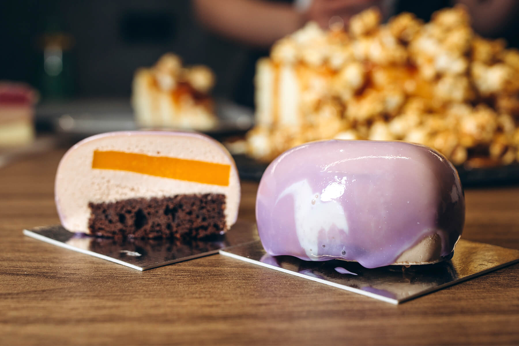 Zwei kleine Mousse-Törtchen auf silbernen Tortenunterlagen, links eines halbiert mit heller Mousse, einer orangegelben Einlage und dunklem Schokoladenbiskuit als Boden, rechts ein ganzes mit lila Spiegelglasur und weißem Streifen