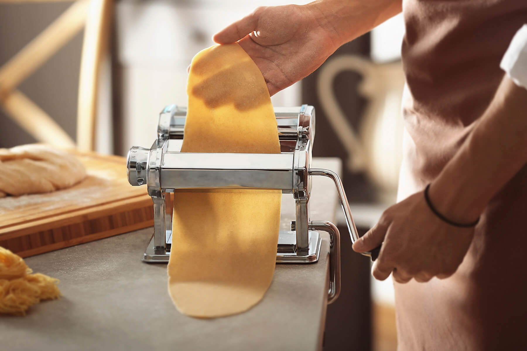 Eine Hand führt eine lange dünne Teigbahn durch die glatten Walzen einer festgeschraubten Nudelmaschine, die andere Hand dreht die Kurbel
