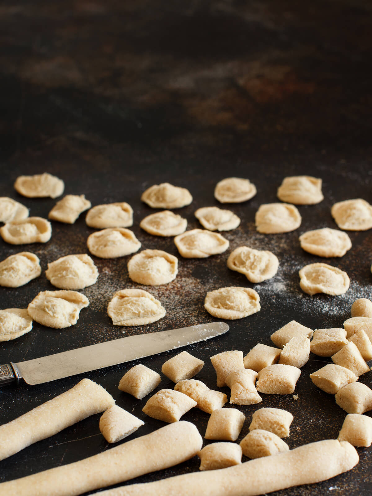 Reihen fertig geformter Orecchiette auf dunkler Arbeitsfläche, davor ein Tafelmesser, geschnittene Teigwürfel und fingerdicke Teigstränge