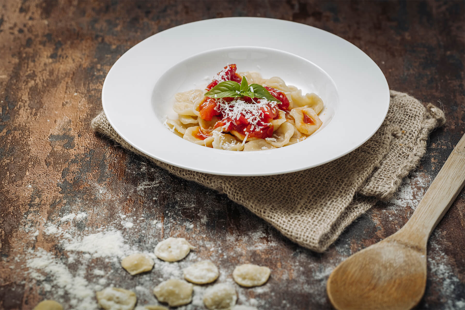 Tiefer weißer Teller mit gekochten Orecchiette in Tomatensauce, geriebenem Käse und Basilikum, davor rohe Öhrchen auf bemehlter Fläche