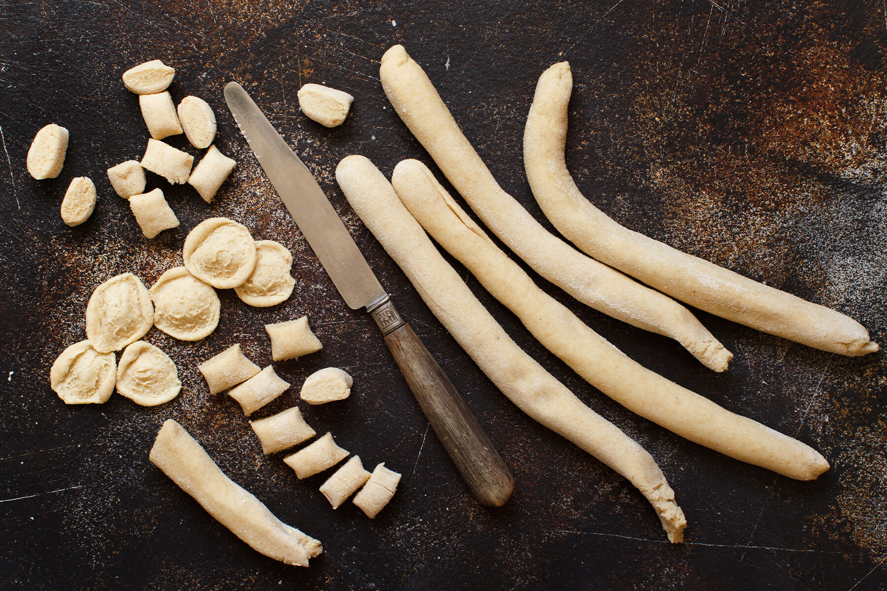 Fingerdicke Teigstränge, daneben abgeschnittene Teigwürfel, aufgezogene Teigmuscheln und fertig geformte Öhrchen sowie ein Tafelmesser mit Holzgriff