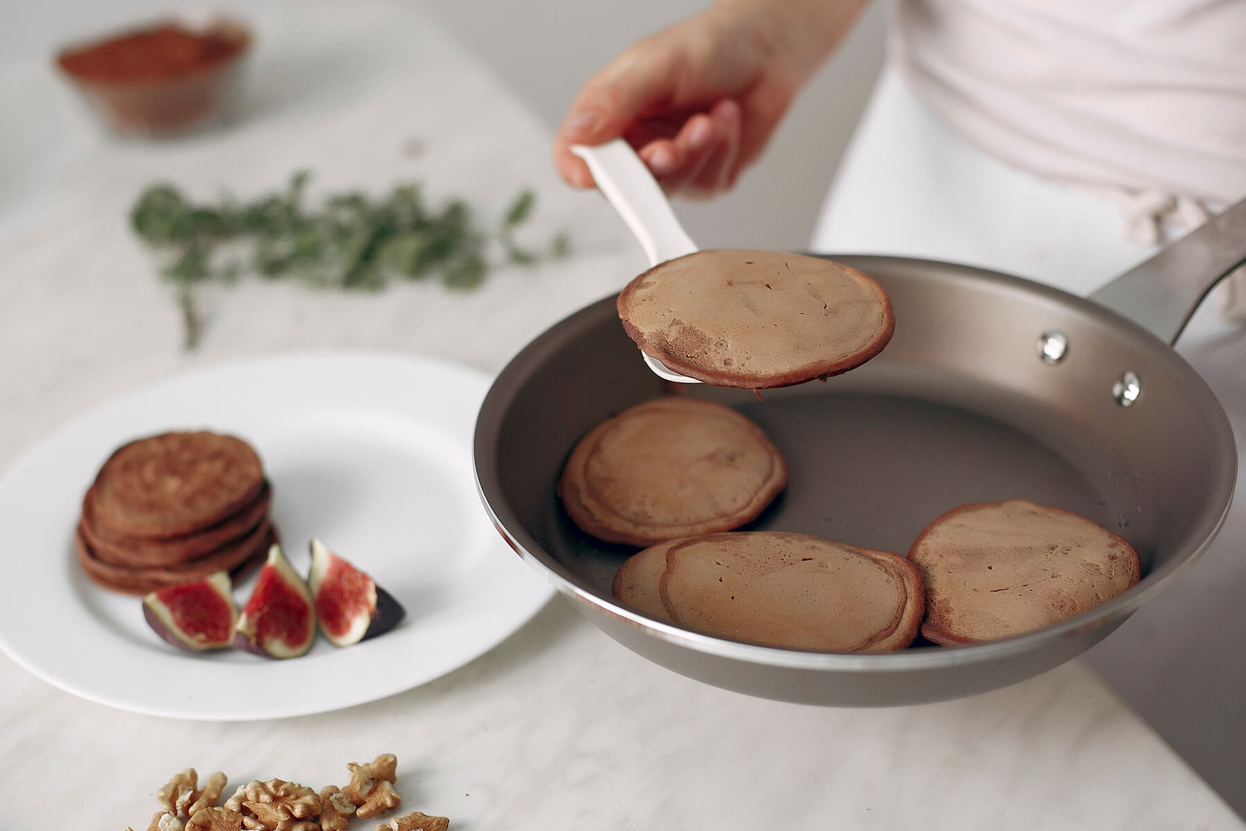 Drei kleine dunkle Pfannkuchen liegen dicht an dicht in einer Pfanne, ein vierter wird mit einem weißen Wender abgehoben