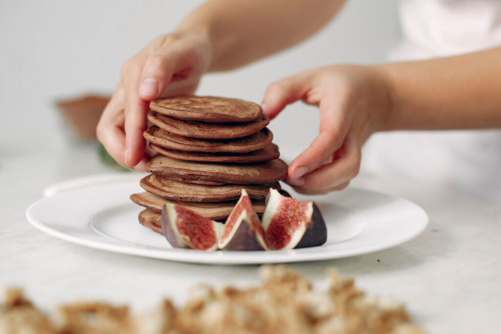 Zwei Hände setzen einen Stapel kleiner dunkler Pfannkuchen auf einen weißen Teller, daneben Feigenviertel