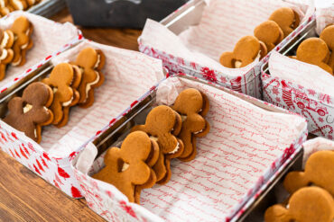 Mehrere rechteckige Blechdosen, mit weihnachtlich bedrucktem Papier ausgelegt, darin Lebkuchen-Männchen mit heller Cremefüllung in Reihen