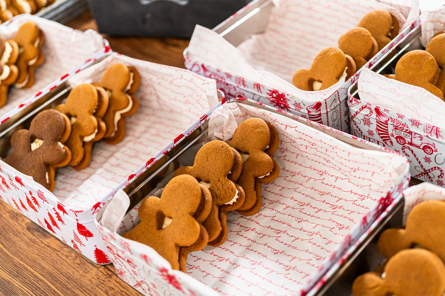 Mehrere rechteckige Blechdosen, mit weihnachtlich bedrucktem Papier ausgelegt, darin Lebkuchen-Männchen mit heller Cremefüllung in Reihen