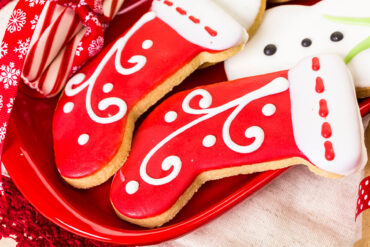 Zwei stiefelförmige Plätzchen mit glatt geflutetem rotem Guss und weiß aufgespritzten Schnörkeln auf einem roten Teller, daneben Zuckerstangen und ein Schneemann-Plätzchen