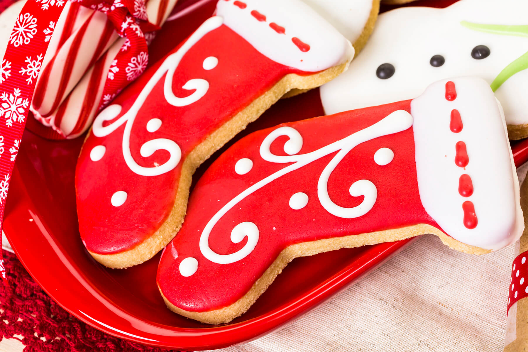Zwei stiefelförmige Plätzchen mit glatt geflutetem rotem Guss und weiß aufgespritzten Schnörkeln auf einem roten Teller, daneben Zuckerstangen und ein Schneemann-Plätzchen