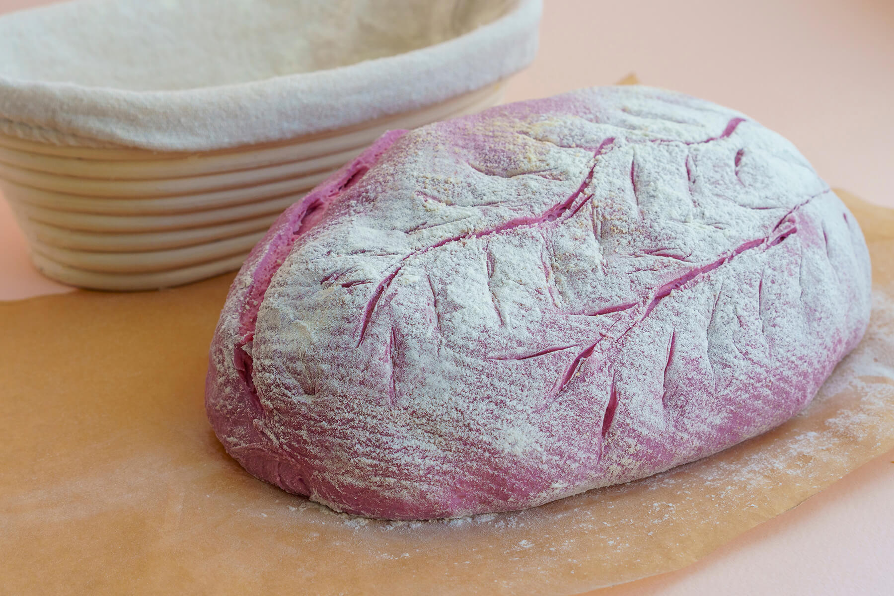 Ovaler, kräftig rosa gefärbter Teigling mit weißer Mehlschicht und eingeschnittenem Blattmuster aus einer Mittellinie mit seitlichen Adern auf Backpapier, dahinter ein Gärkörbchen