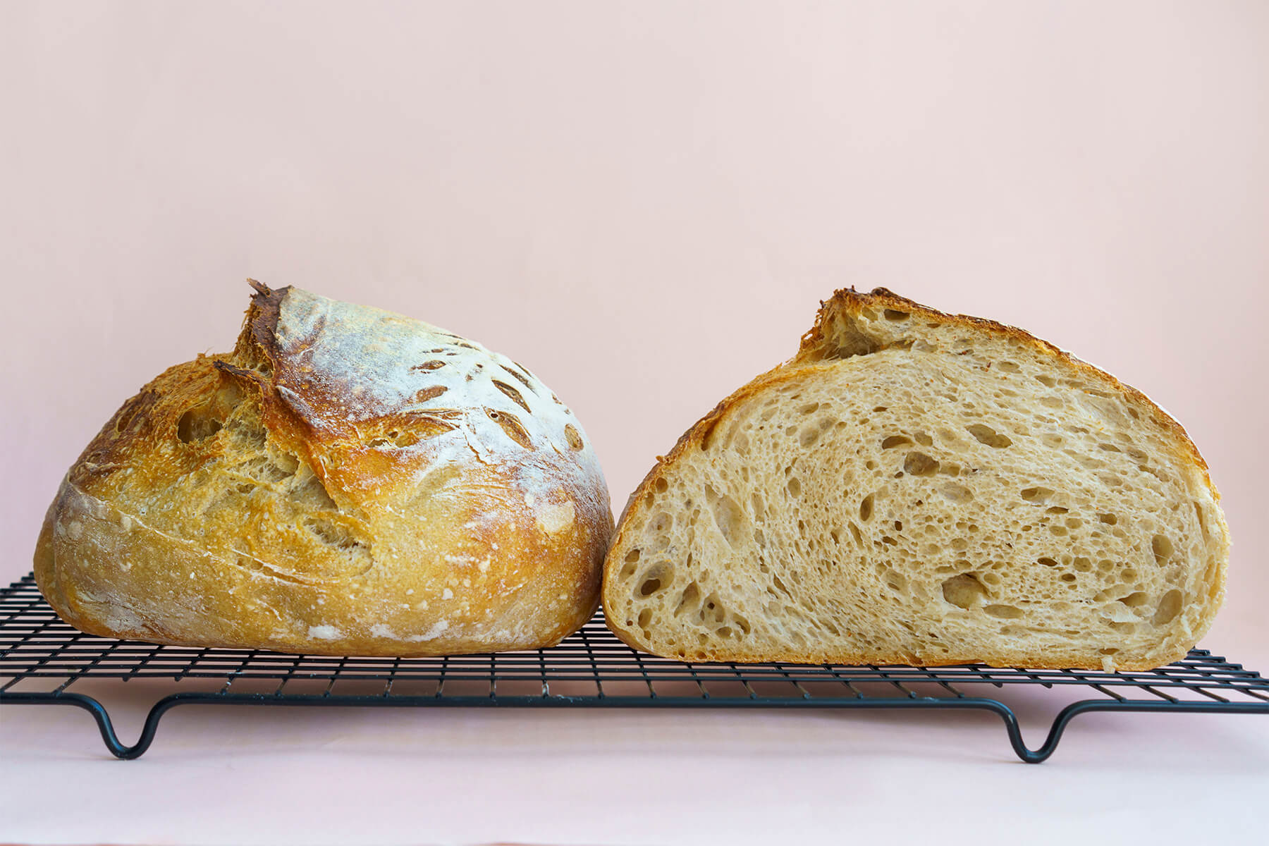 Ein rundes Sauerteigbrot mit aufgestelltem Einschnitt auf einem Kuchengitter, daneben die angeschnittene Hälfte mit offener, unregelmäßiger Krume