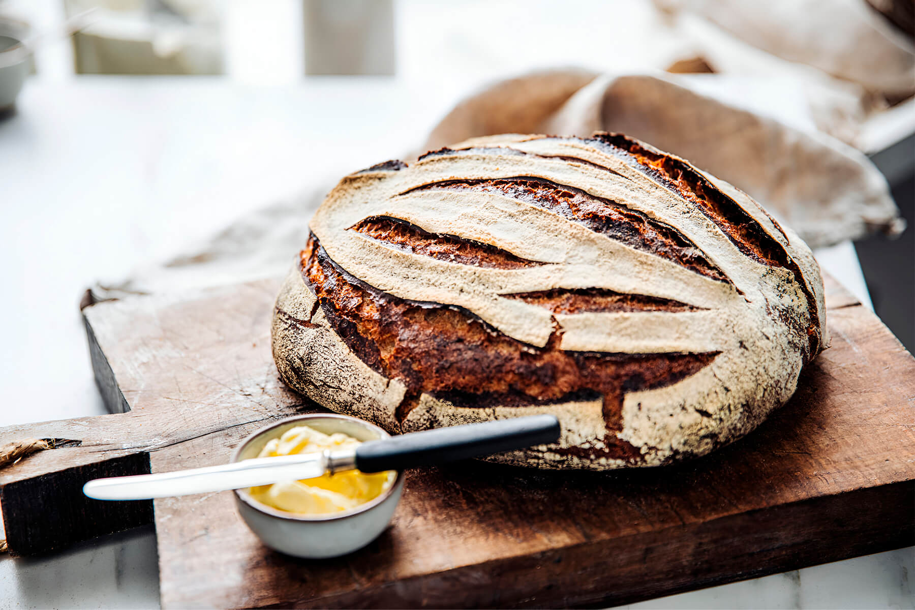 Rundes Sauerteigbrot mit heller Mehlschicht und weit geöffneten, dunkelbraunen Schnitten auf einem Holzbrett, davor ein Schälchen Butter mit einem Messer