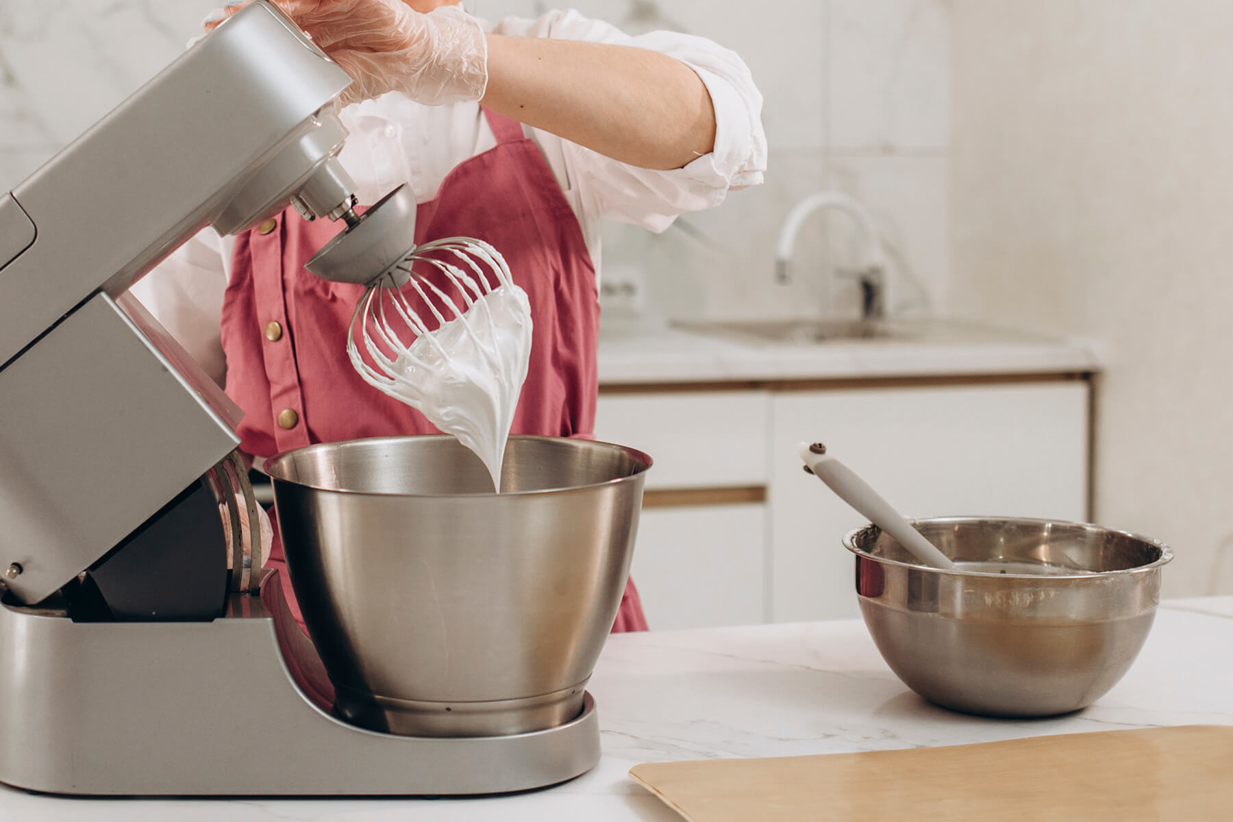 Der abgehobene Schneebesen einer Küchenmaschine hält eine weiße, glänzende aufgeschlagene Masse, darunter steht die Rührschüssel