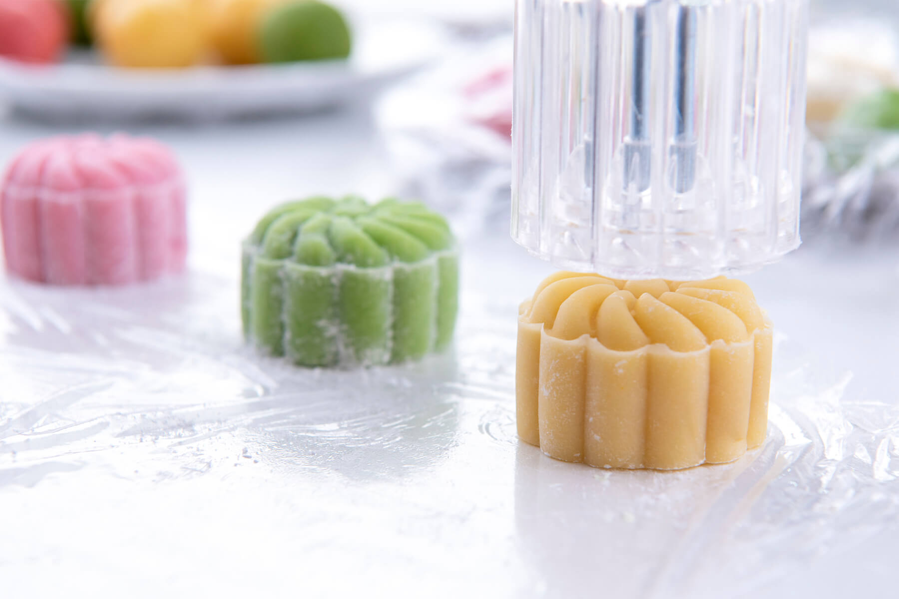 Durchsichtige Stempelform wird von einem gelben Mondkuchen mit Blütenmuster und geriffeltem Rand abgehoben, links dahinter ein grüner und ein rosa Mondkuchen auf Frischhaltefolie