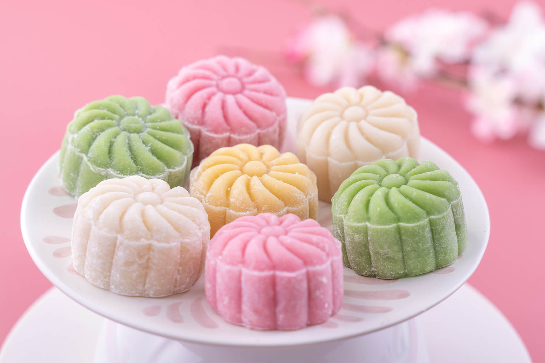 Sieben gepresste Mondkuchen mit Blütenmuster auf einem weißen Tortenständer vor rosa Hintergrund, je zwei in Grün, Rosa und Weiß und einer in Gelb, dahinter unscharfe blühende Zweige