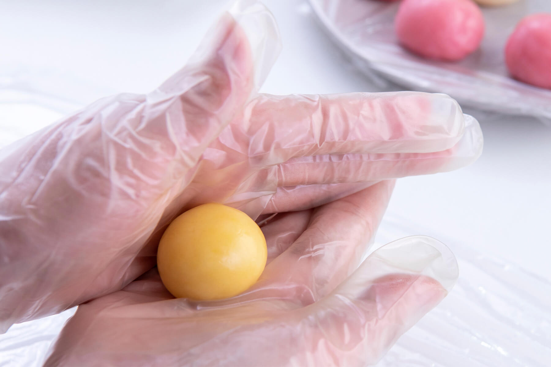 Glatte gelbe Teigkugel zwischen zwei Händen in Einweghandschuhen, im Hintergrund rosa Teigkugeln auf einem mit Folie bespannten Teller