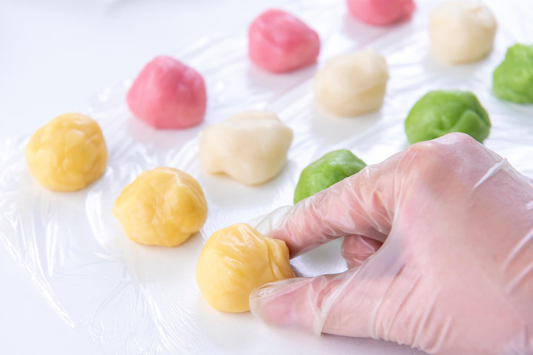 Zwölf ungeformte Teigportionen in Gelb, Weiß, Rosa und Grün liegen in Reihen auf Frischhaltefolie, eine Hand im Einweghandschuh fasst die vordere gelbe Portion an