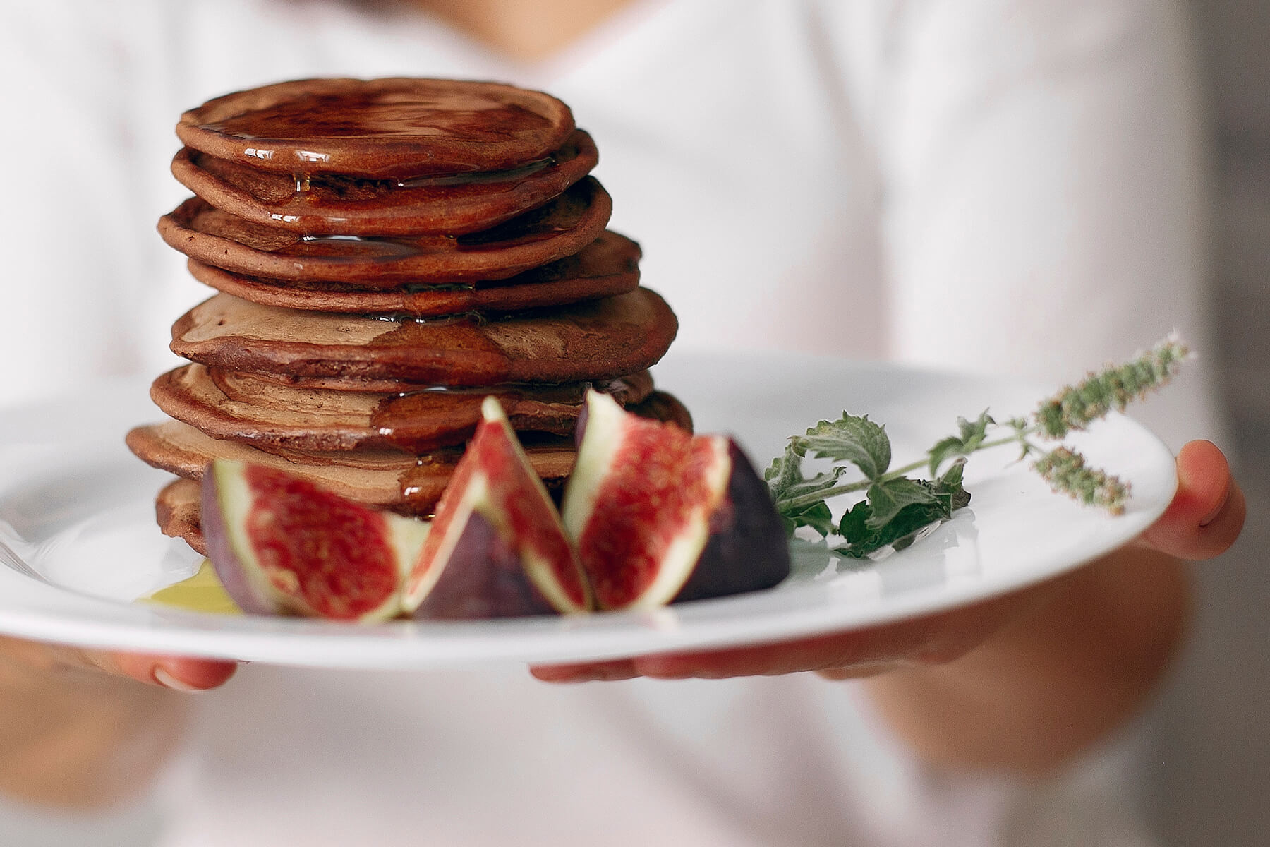 Ein hoher Stapel kleiner Schokoladen-Pfannkuchen mit Honig auf einem weißen Teller, daneben Feigenviertel und ein Minzzweig