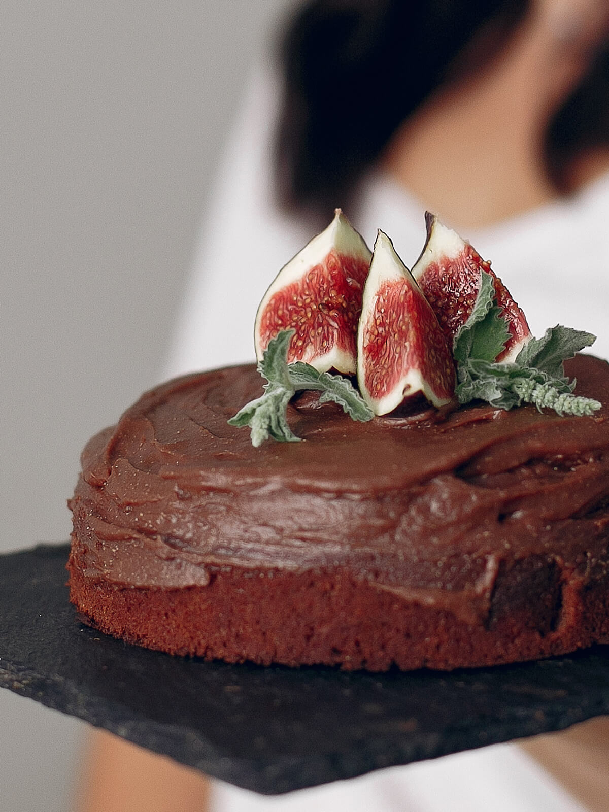 Eine kleine Schokoladentorte mit gewölbter Oberseite und dick aufgestrichener Schokoladencreme auf einer Schieferplatte, darauf drei aufrecht stehende Feigenviertel und ein paar Kräuterblätter