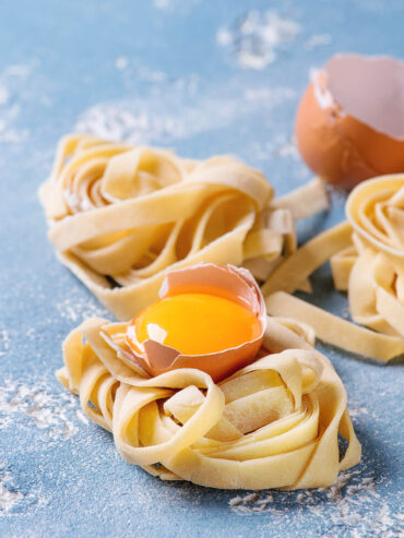 Drei frisch gedrehte Tagliatelle-Nester auf bemehlter blauer Fläche, auf dem vorderen Nest liegt ein Eigelb in der halben Schale
