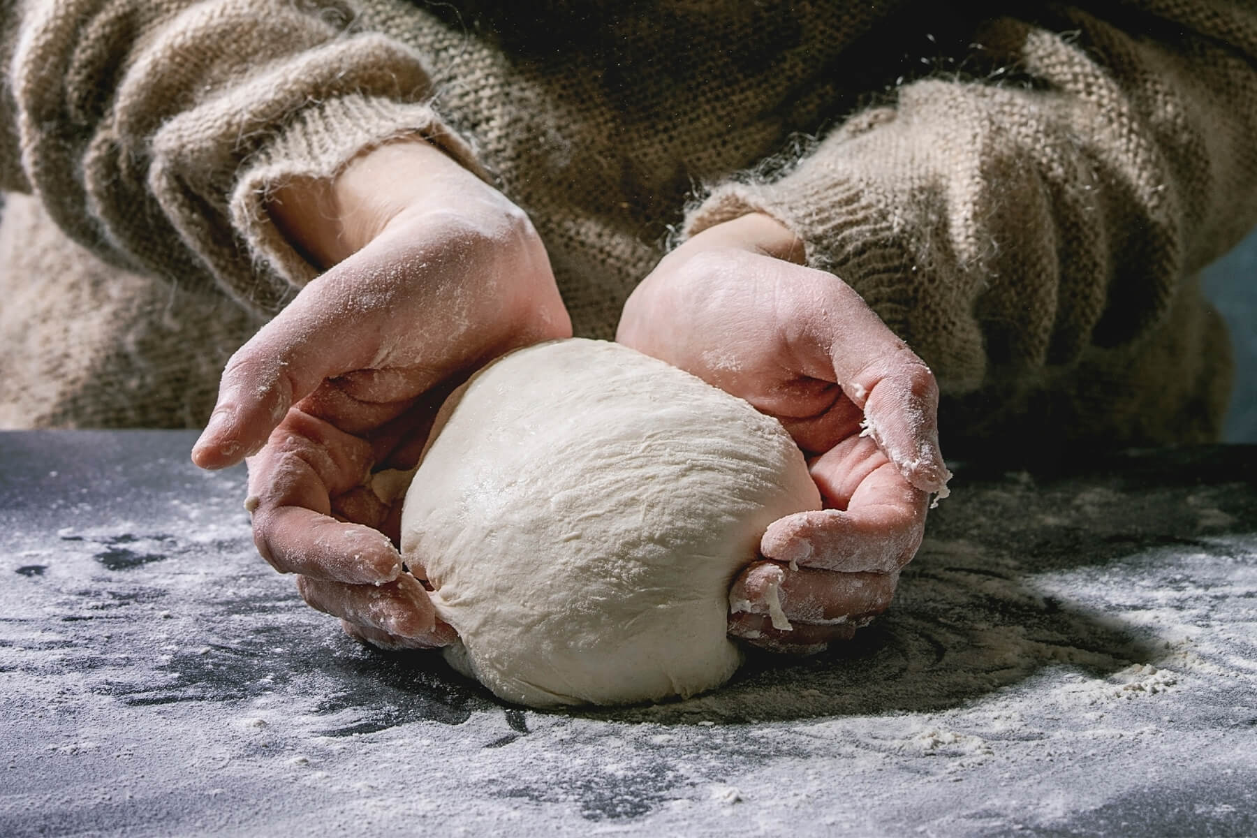 Zwei bemehlte Hände umfassen einen hellen Brotteig auf einer dunklen, bemehlten Arbeitsfläche und formen ihn zu einer runden Kugel