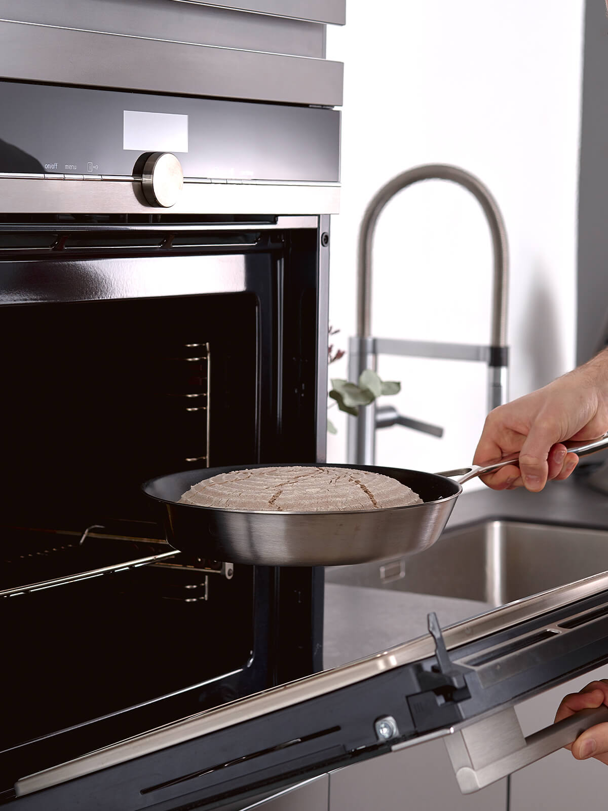 Eine Hand schiebt eine Edelstahlpfanne mit Metallgriff und einem bemehlten, eingeschnittenen Teigling in einen geöffneten Backofen