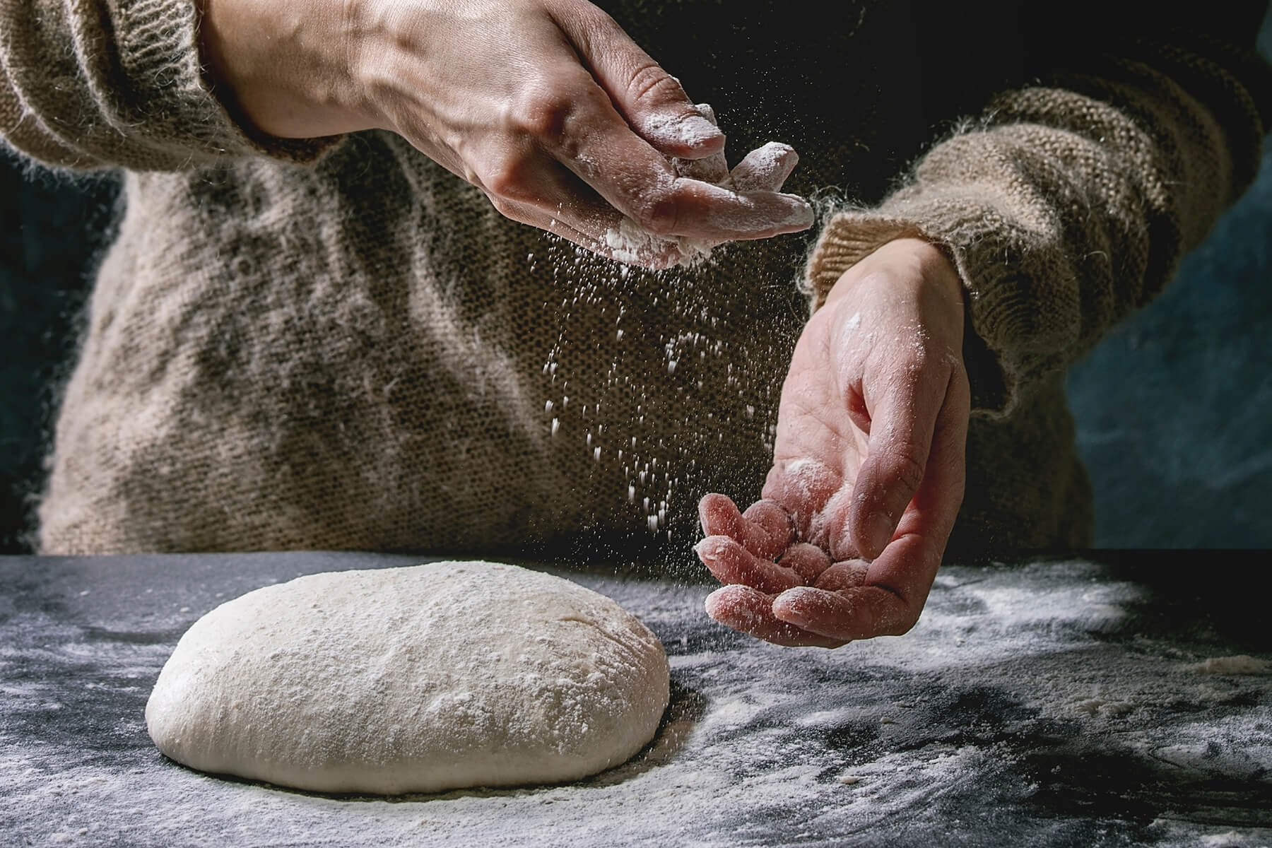 Eine Hand lässt Mehl auf einen rund gewirkten Teigling auf einer dunklen Arbeitsfläche rieseln