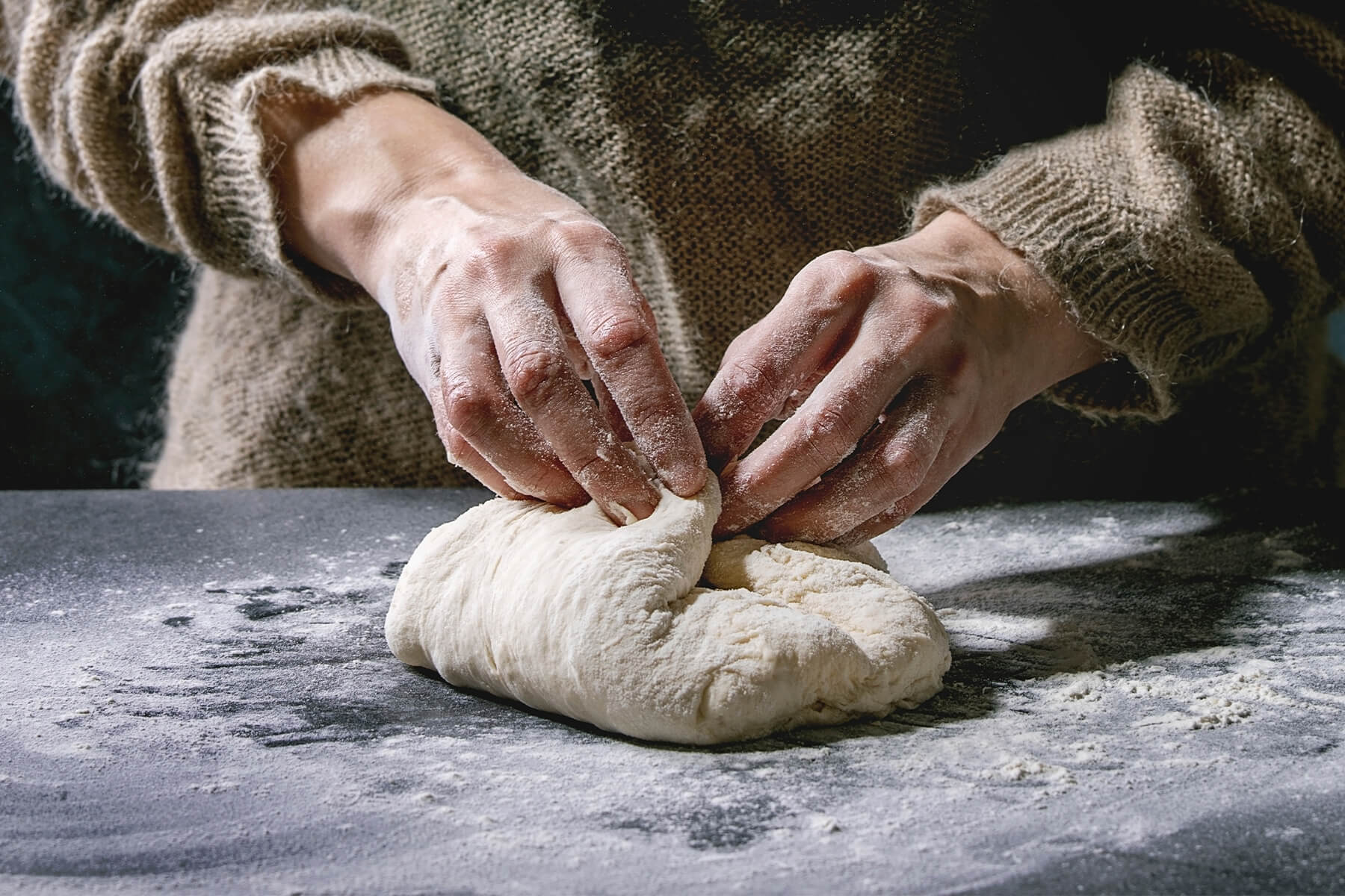 Zwei bemehlte Hände ziehen die Ränder eines hellen Brotteigs auf einer dunklen, bemehlten Arbeitsfläche zur Mitte