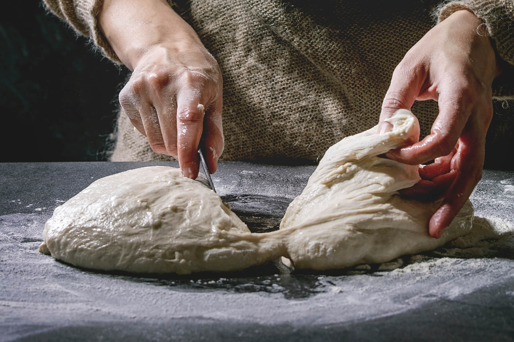 Zwei Teigstücke auf einer dunklen Arbeitsfläche, zwischen denen sich beim Teilen dünne Teigfäden spannen, eine Hand hält ein Messer