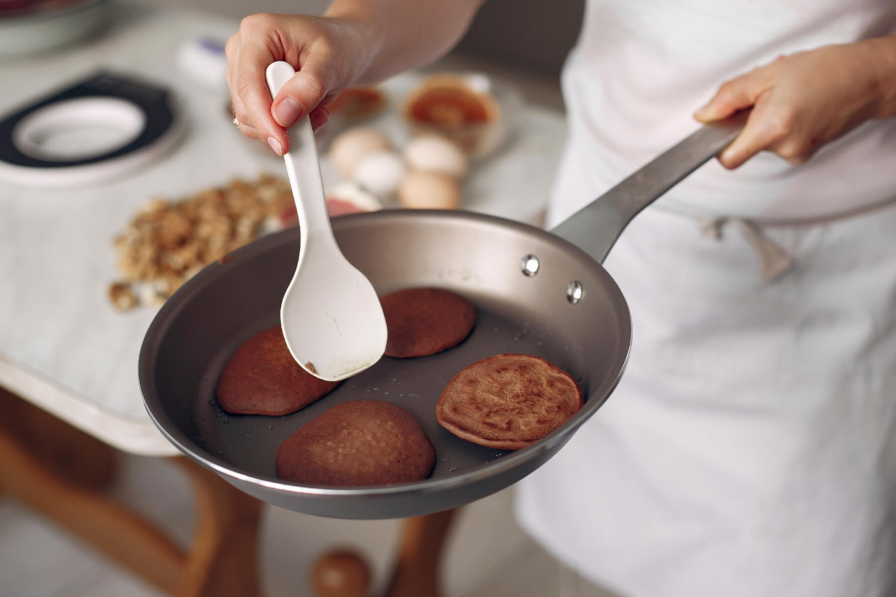 Vier kleine dunkle Pfannkuchen in einer beschichteten Pfanne, einer davon bereits gewendet und goldbraun gefleckt, daneben ein weißer Wender