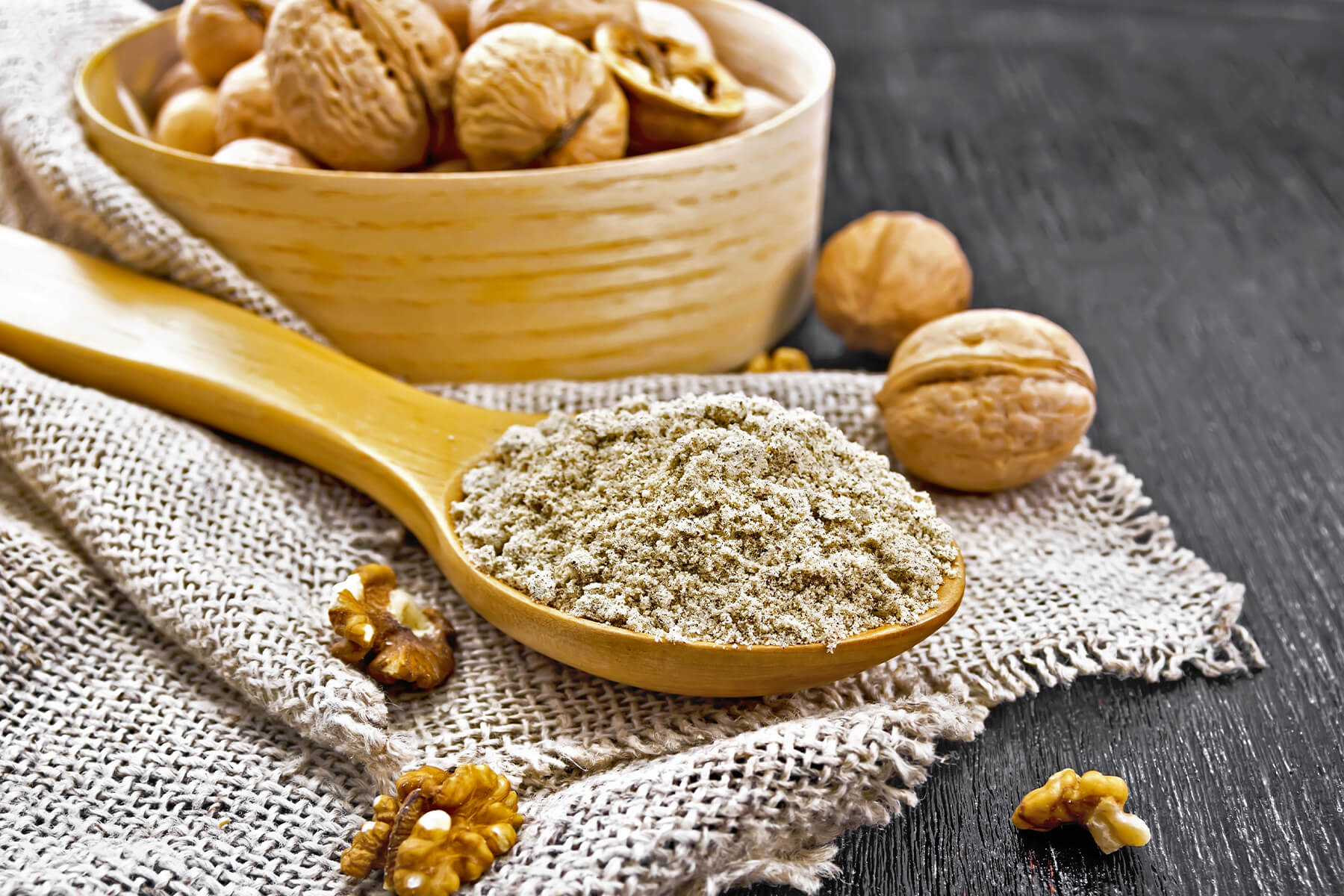 Holzlöffel mit feinem, graubeigem Walnussmehl voller dunkler Pünktchen auf Sackleinen über dunklem Holz, dahinter eine helle Holzschale mit Walnüssen in der Schale, daneben zwei ganze Walnüsse und einzelne Walnusskerne