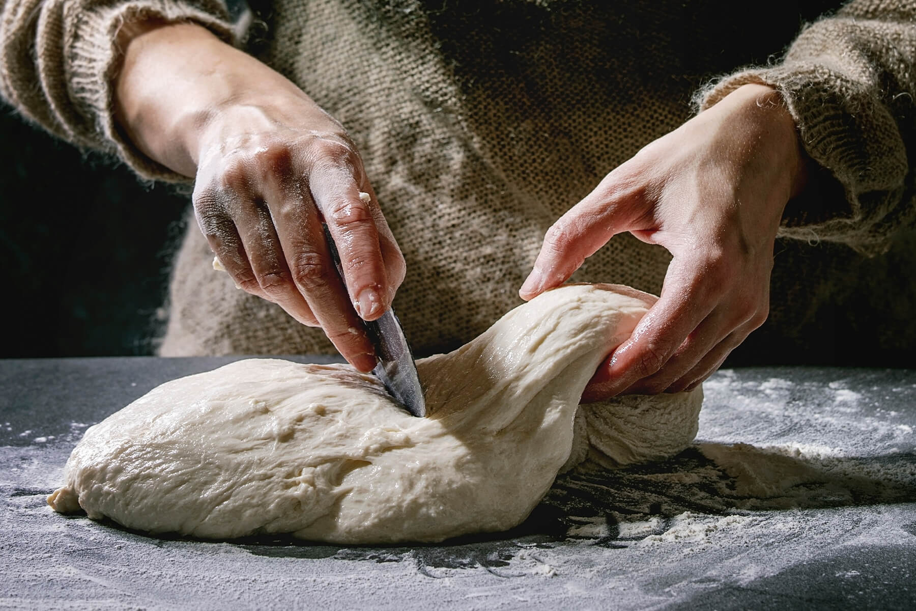 Eine Hand teilt einen weichen Brotteig auf einer dunklen, bemehlten Arbeitsfläche mit einem Messer, die andere hält das abgetrennte Stück