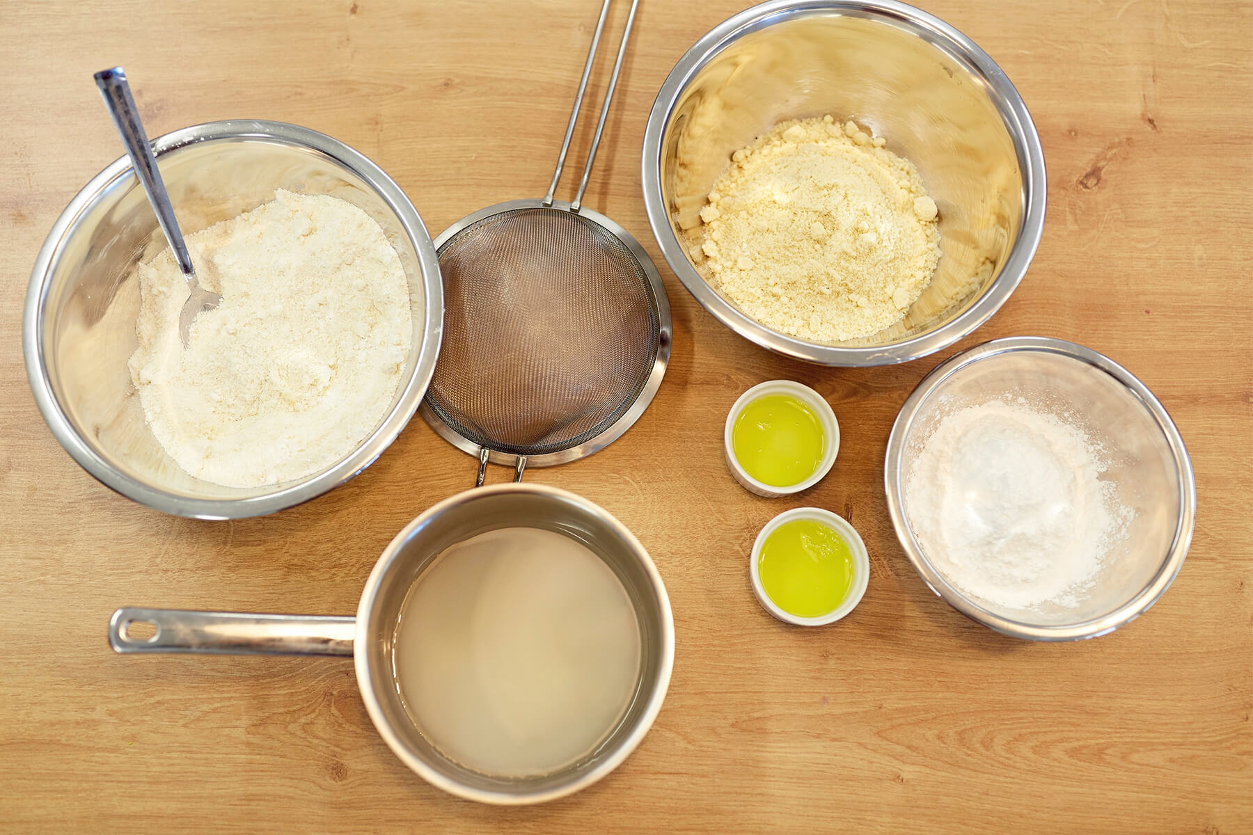 Von oben aufgenommene Arbeitsfläche mit einer Schüssel Baisermasse, einem feinen Sieb, gemahlenen Mandeln, zwei kleinen Schalen Eiweiß, einem Topf mit klarer Zuckerlösung und einer Schüssel Puderzucker