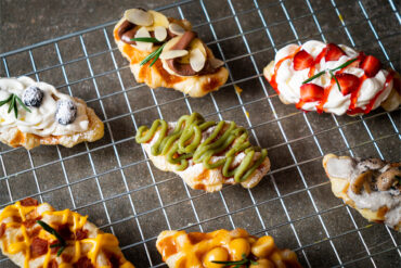 Croffles mit verschiedenen Belägen auf einem Kuchengitter: Matcha-Creme, Schokolade mit Banane und Mandelblättchen, Sahne mit Erdbeeren, Sahne mit Heidelbeeren, Champignons in Rahmsauce, Käsesauce mit Speck und Karamell mit Macadamia
