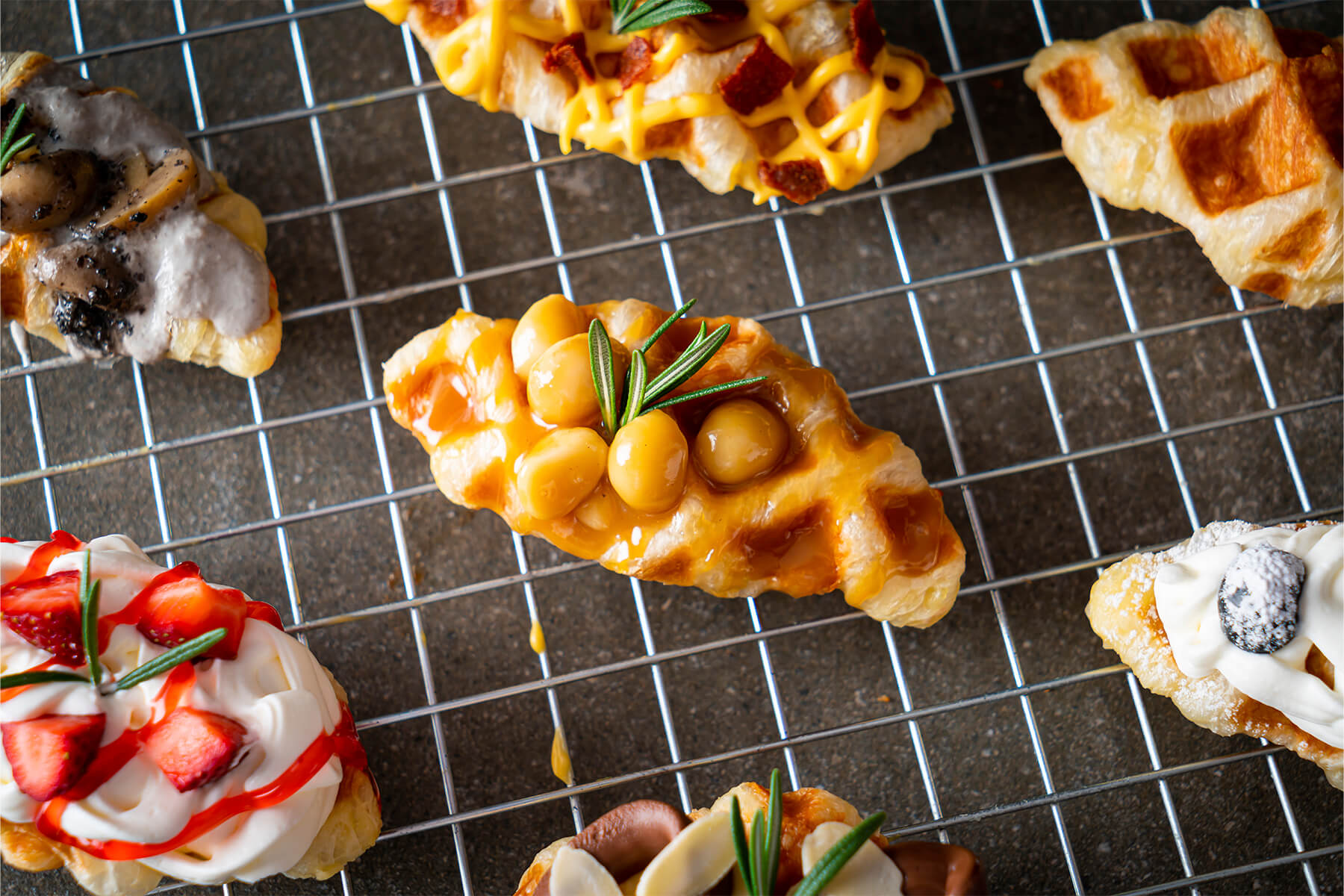 Croffle mit glänzender Karamellsauce und ganzen Macadamianüssen auf einem Kuchengitter, oben rechts ein Croffle ohne Belag mit goldbraunen Waffelfeldern