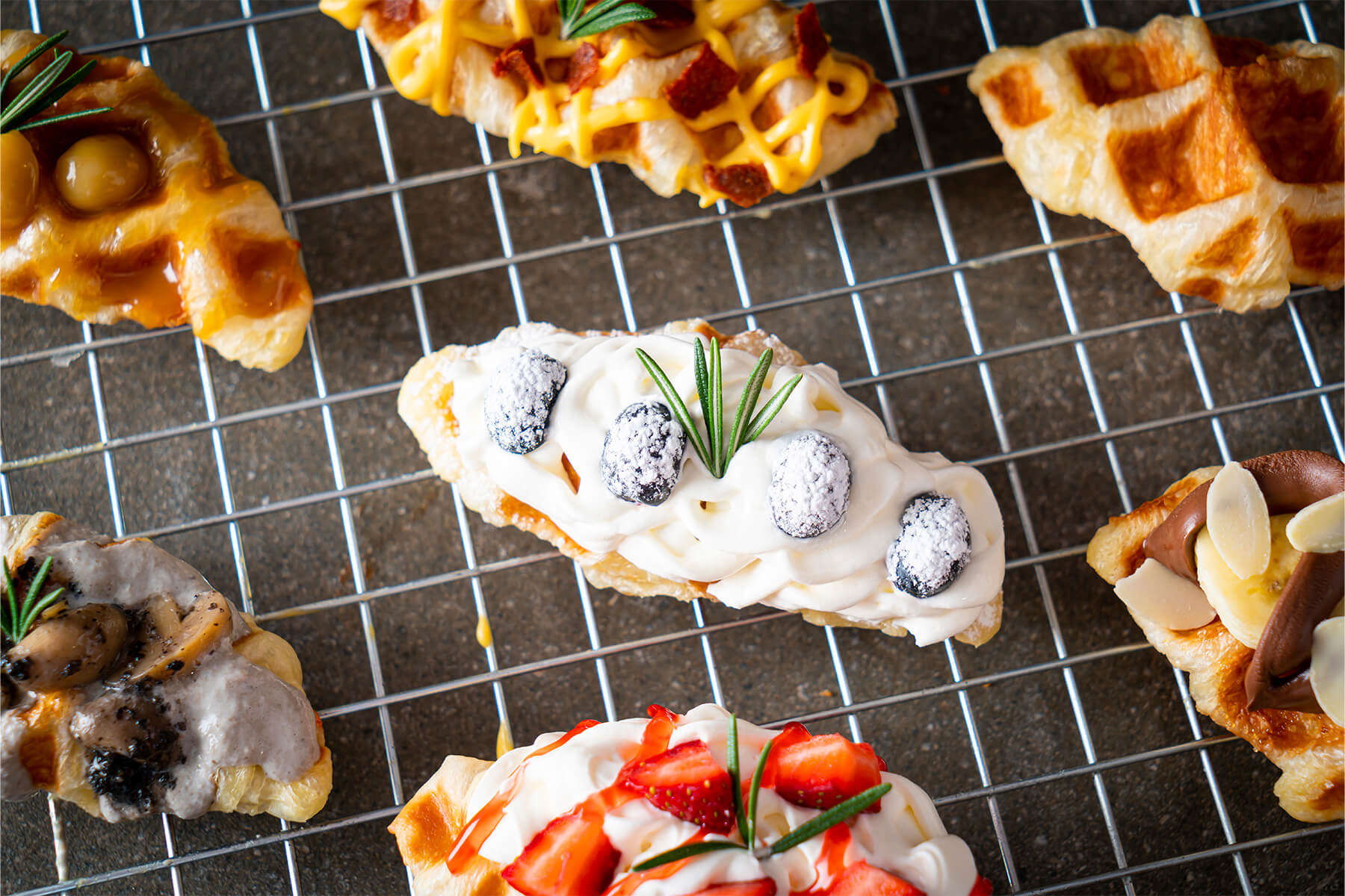 Croffle mit Schlagsahne, vier mit Puderzucker bestäubten Heidelbeeren und einem Rosmarinzweig auf einem Kuchengitter, darum weitere Croffles mit verschiedenen Belägen