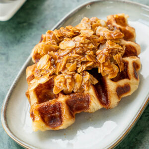 Croissant aus dem Waffeleisen mit glänzender Karamellkruste im Waffelmuster, darauf ein Berg Mandelblättchen in Karamell, auf einem ovalen Keramikteller