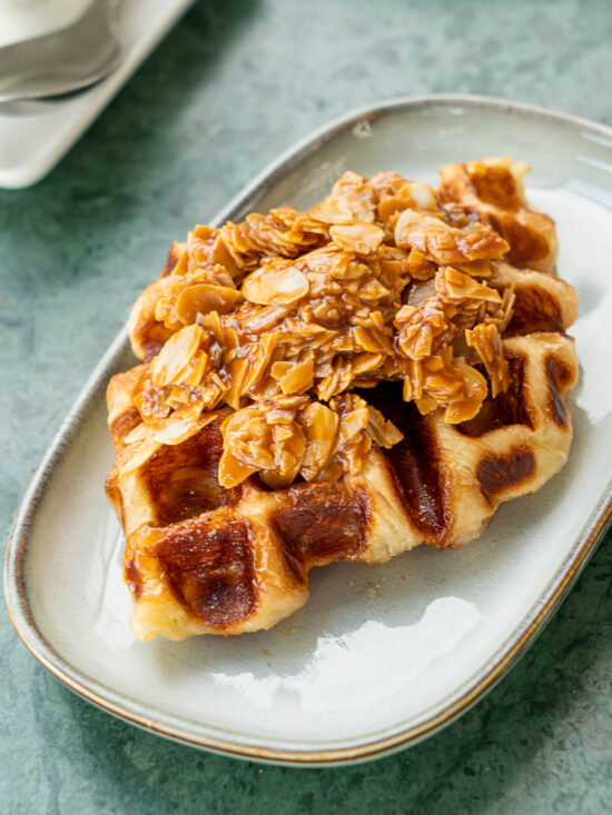 Croissant aus dem Waffeleisen mit glänzender Karamellkruste im Waffelmuster, darauf ein Berg Mandelblättchen in Karamell, auf einem ovalen Keramikteller