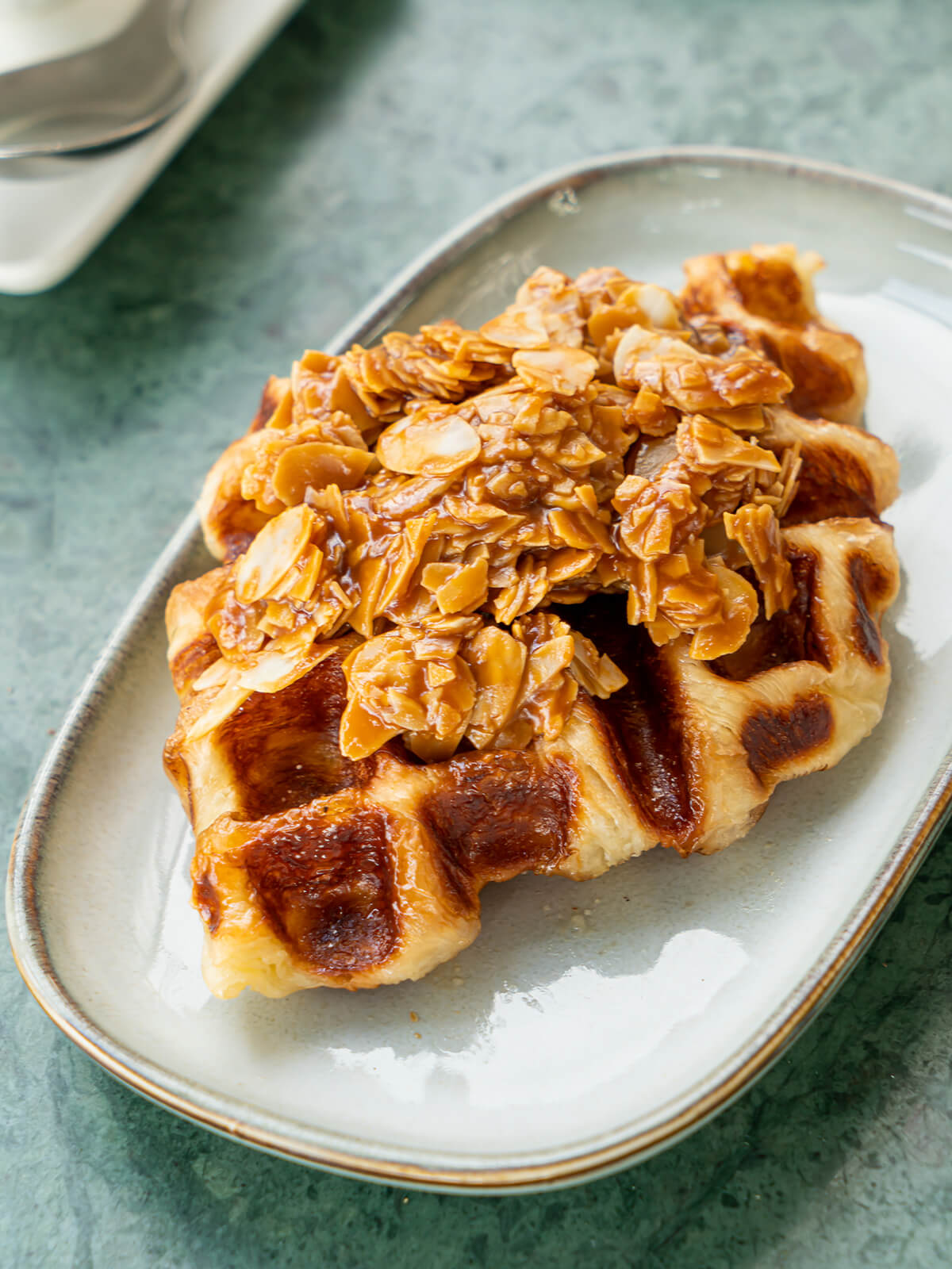 Croissant aus dem Waffeleisen mit glänzender Karamellkruste im Waffelmuster, darauf ein Berg Mandelblättchen in Karamell, auf einem ovalen Keramikteller