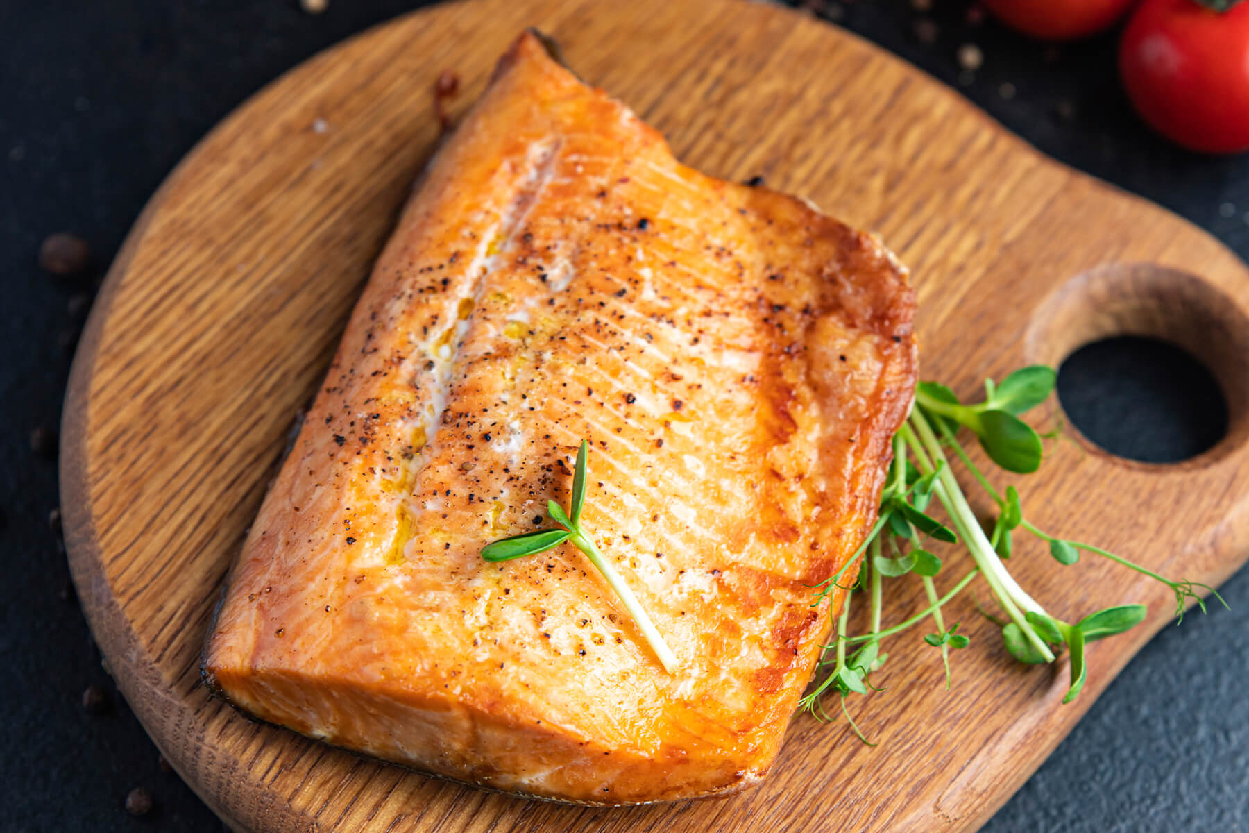 Gebackenes Lachsfilet auf einem Holzbrett, daneben ein Büschel Sprossen, im Hintergrund Kirschtomaten