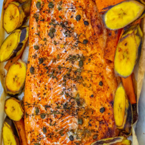Gebackene Lachsseite mit Thymianzweigen, Kapern und Paprika auf Backpapier, ringsum gebackene Kochbananenscheiben mit Schale und Möhrenstifte