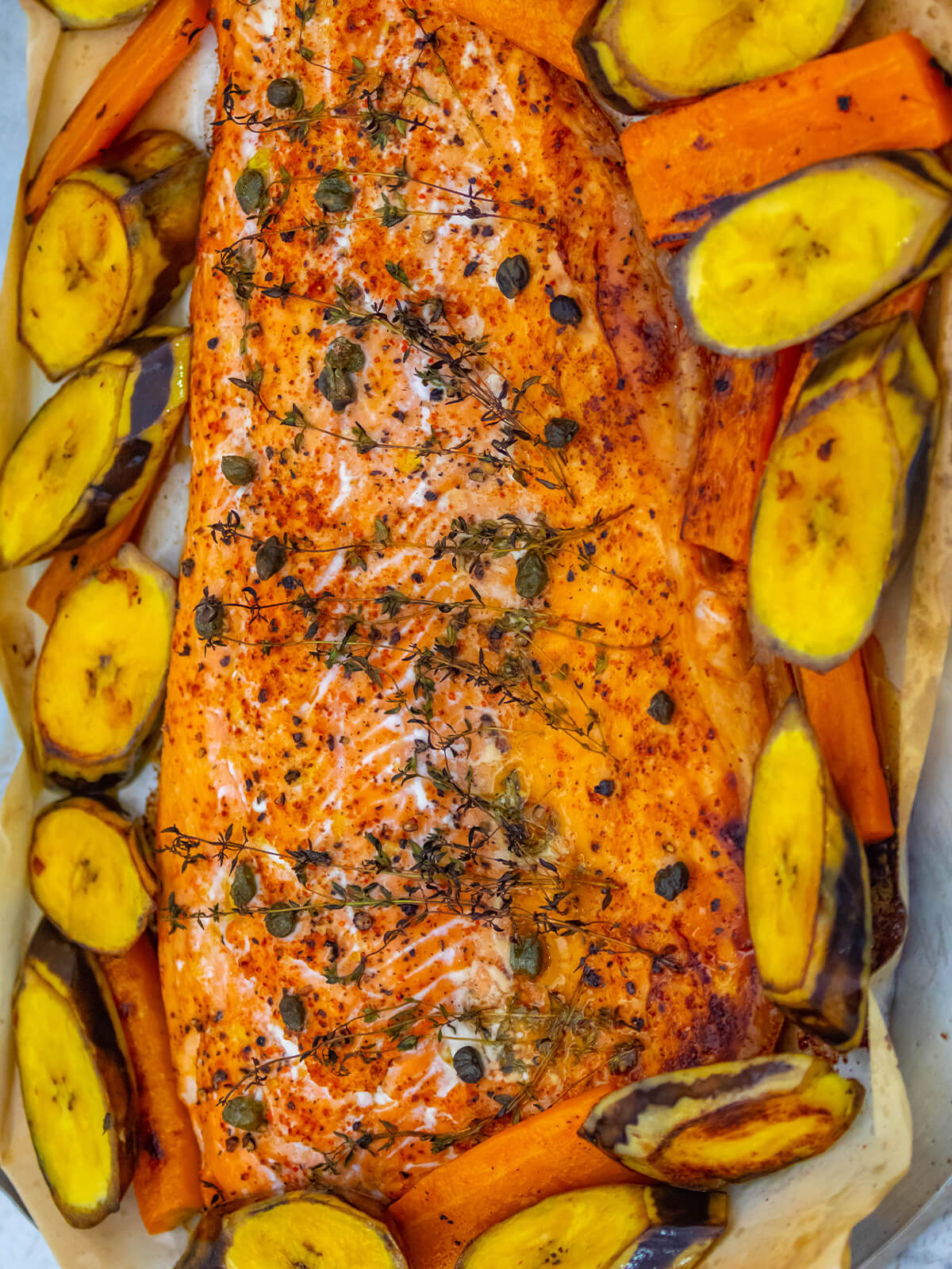Gebackene Lachsseite mit Thymianzweigen, Kapern und Paprika auf Backpapier, ringsum gebackene Kochbananenscheiben mit Schale und Möhrenstifte