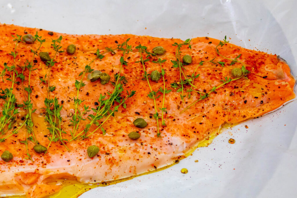 Rohe, mit Paprika und Pfeffer gewürzte Lachsseite mit Thymianzweigen und Kapern auf Backpapier, am Rand Olivenöl
