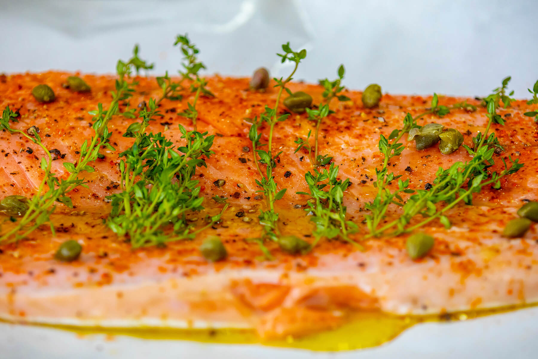 Rohe Lachsseite mit Paprika, grobem Pfeffer, Thymianzweigen und Kapern, darunter Olivenöl auf Backpapier