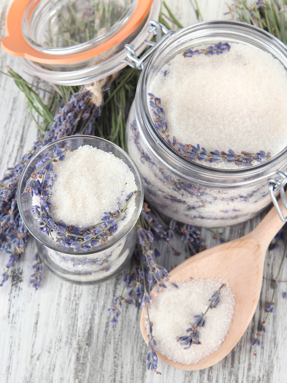 Ein Bügelglas und ein kleineres Glas mit weißem Zucker, an der Glaswand ganze getrocknete Lavendelähren, davor ein Holzlöffel mit Zucker und einer Ähre, ringsum Sträuße getrockneter Lavendel auf hellem Holz