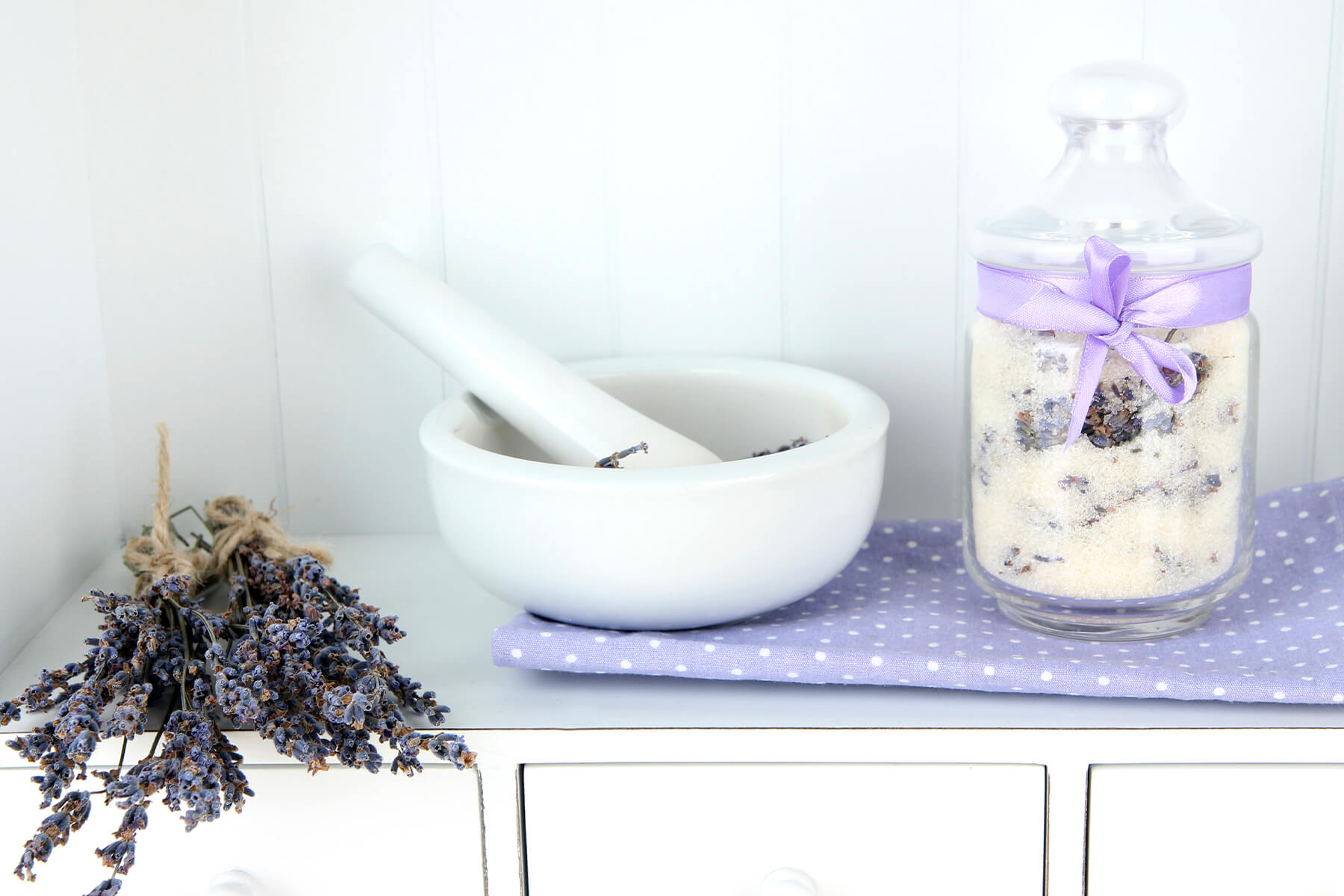 Weißer Mörser mit Stößel und ein paar Lavendelblüten, daneben ein Glas Lavendelzucker mit fliederfarbener Schleife auf einem gepunkteten Tuch, links ein Bund getrockneter Lavendel auf einer weißen Kommode